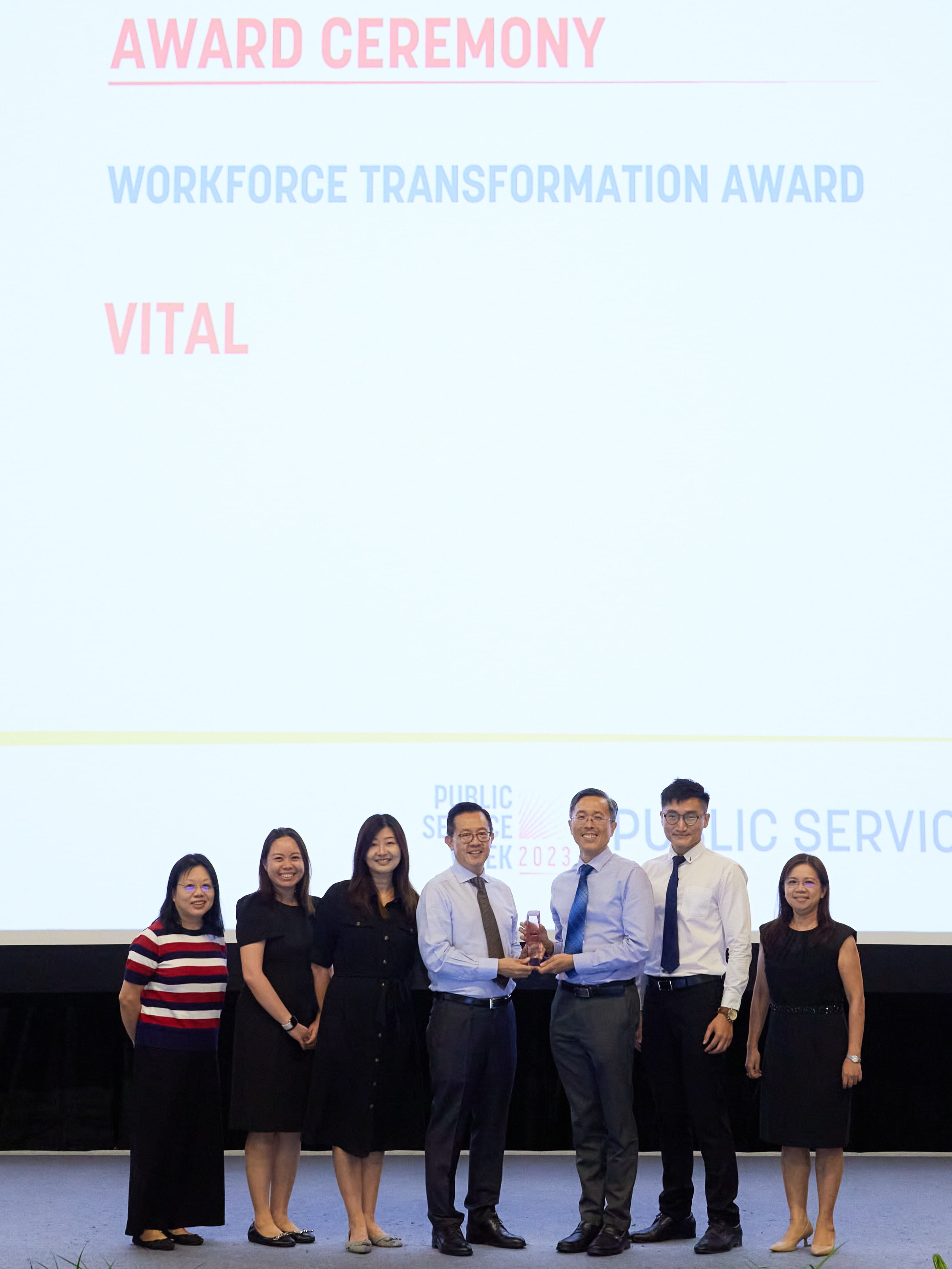Seven people stand on a stage presenting the Workforce Transformation Award to VITAL at a 2023 Public Service Week ceremony.